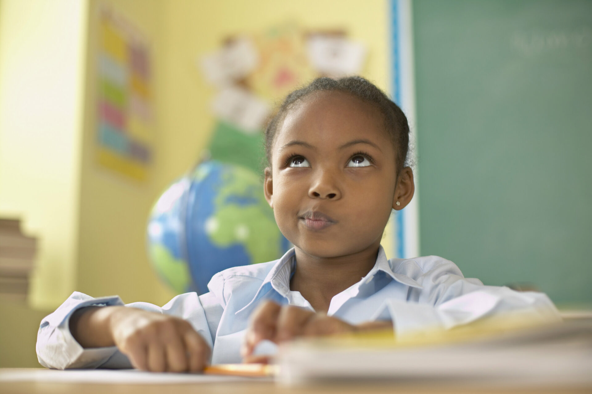 Aluna com cara de pensativa em uma sala de aula, sentada na carteira com papel e lápis na mão. Ela é negra, tem por volta de 6 anos e veste uma camisa azul claro.
